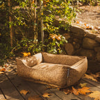 Patterned dog bed on a wooden deck with autumn leaves and a stone wall in the background